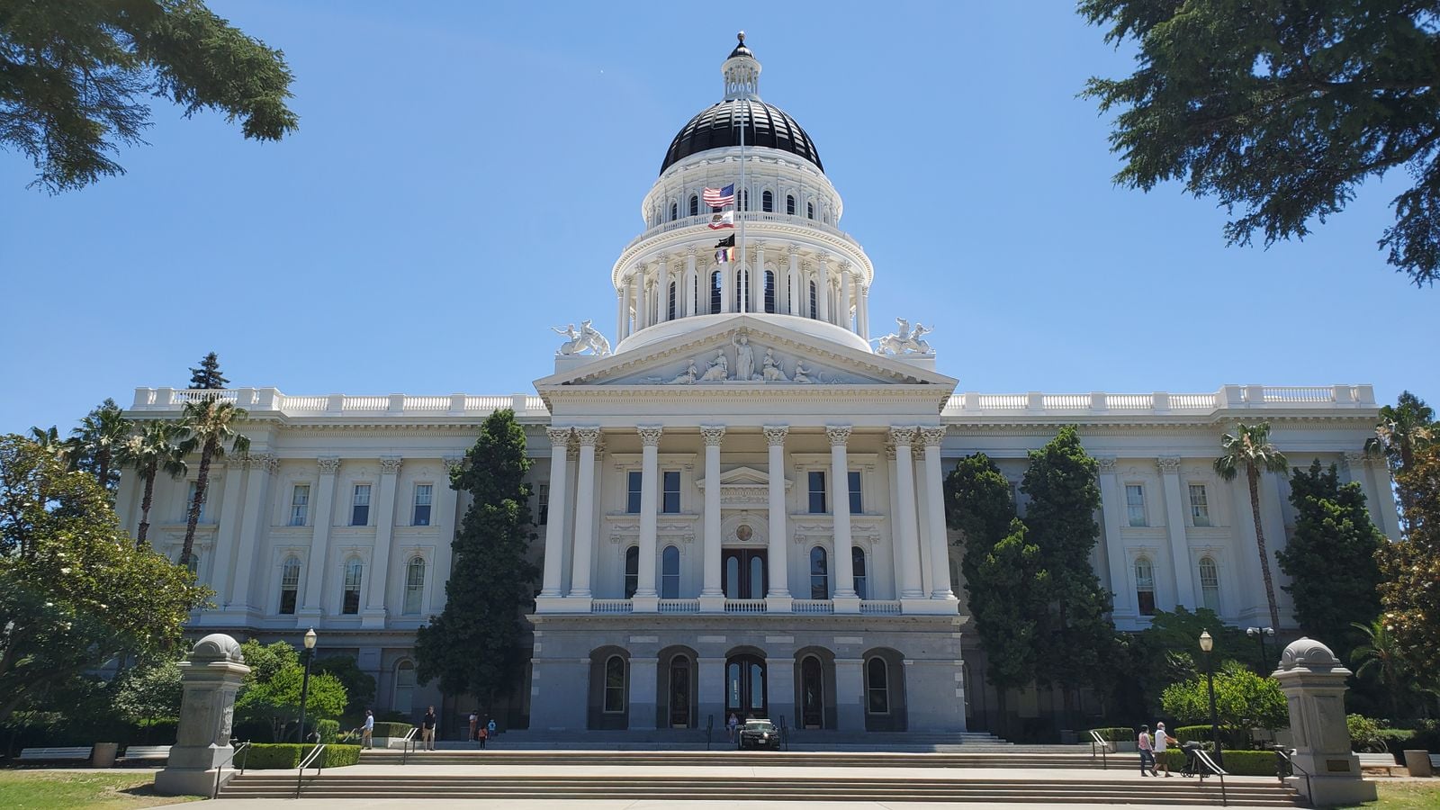 White capitol building with a large domed roof, columns, and flags against a clear blue sky.